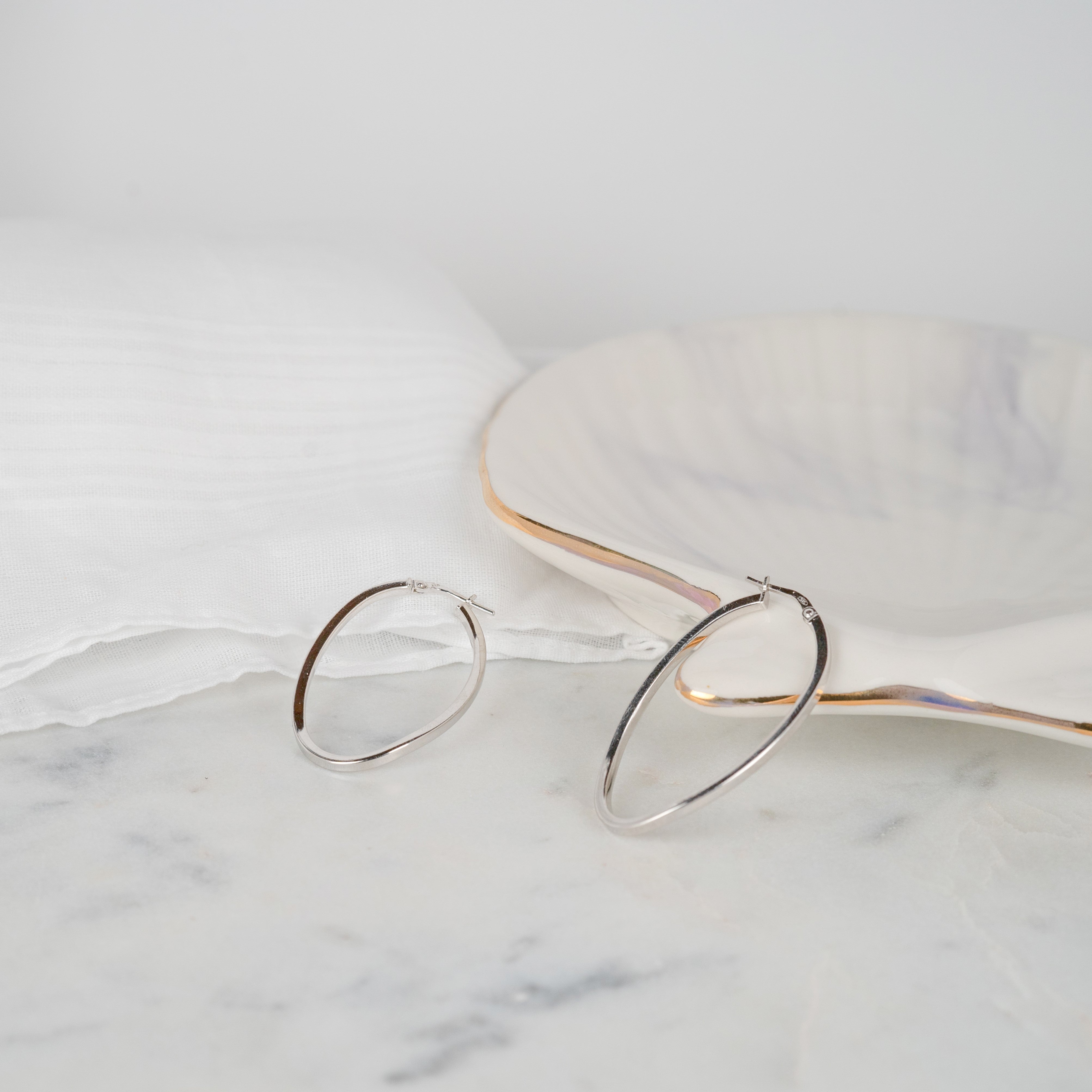 Boucles d'oreilles grandes ovales couleur argent posée sur du marbre et un coquillage